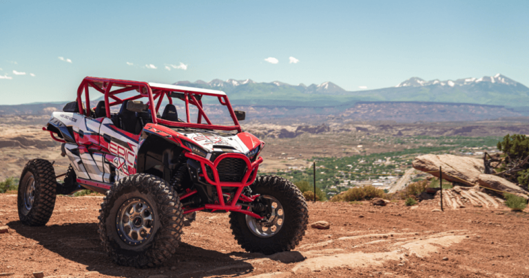 A red and white UTV rests on a dirt path, set against a backdrop of trees and foliage.