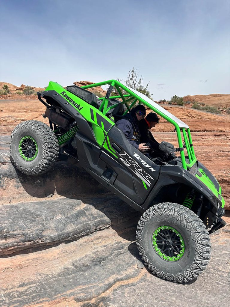 A green and black UTV navigates a rocky hill, Poison Spider showcasing off-road capabilities.