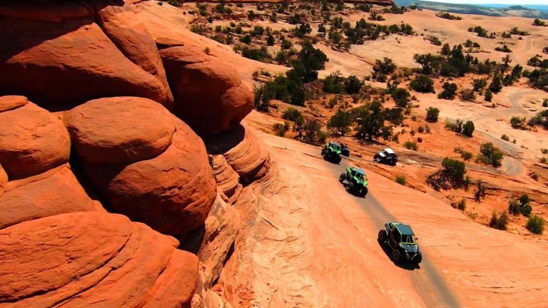 Four vehicles travel along a picturesque road flanked by vibrant red rock cliffs.